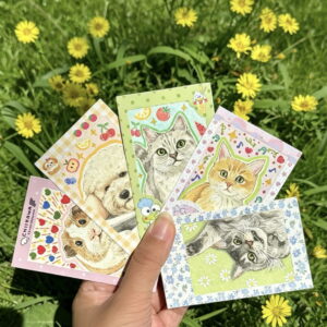 Hand holding mini pet portrait cards over grass and flowers